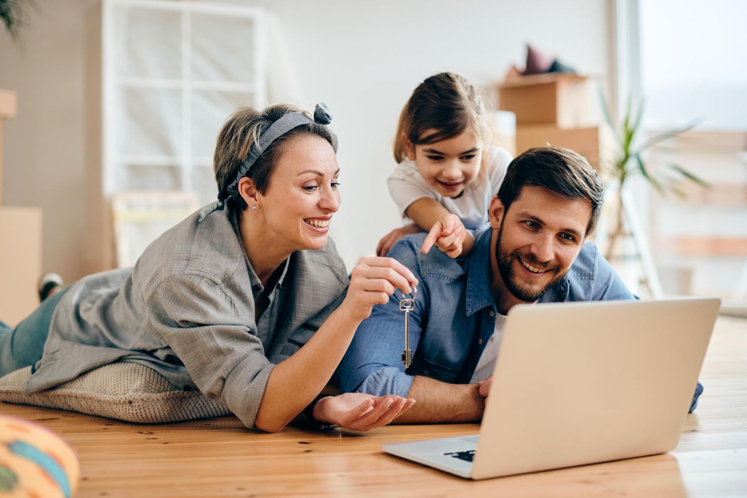 Family on floor looking at laptop, holding keys, child pointing, surrounded by boxes.