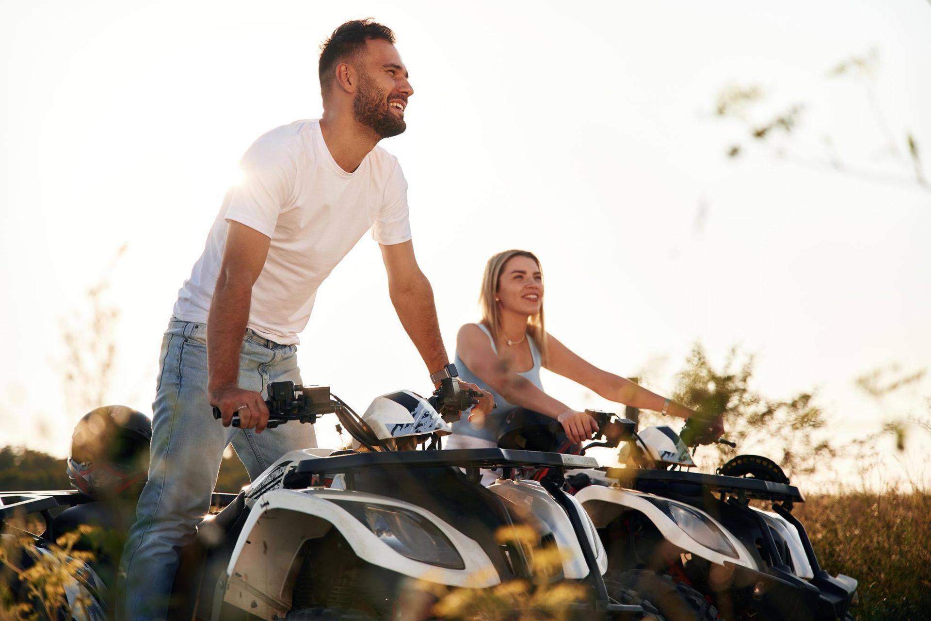 Man and woman ride ATVs outdoors, smiling. Bright sunlight.