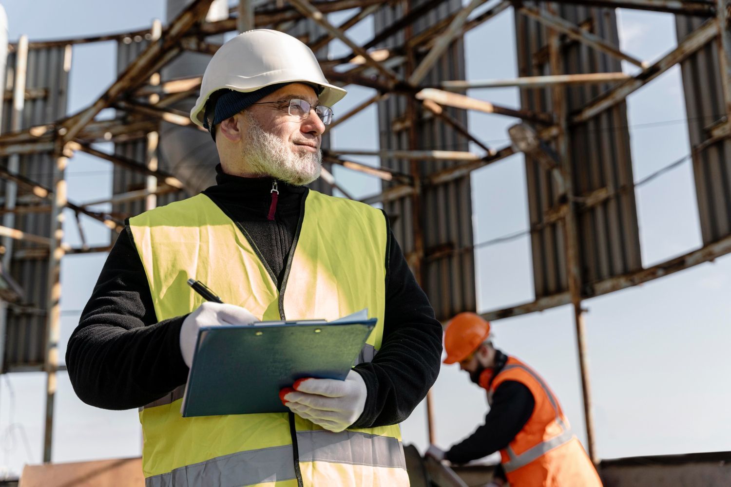 Construction worker in hard hat and vest taking notes with another worker in the background.