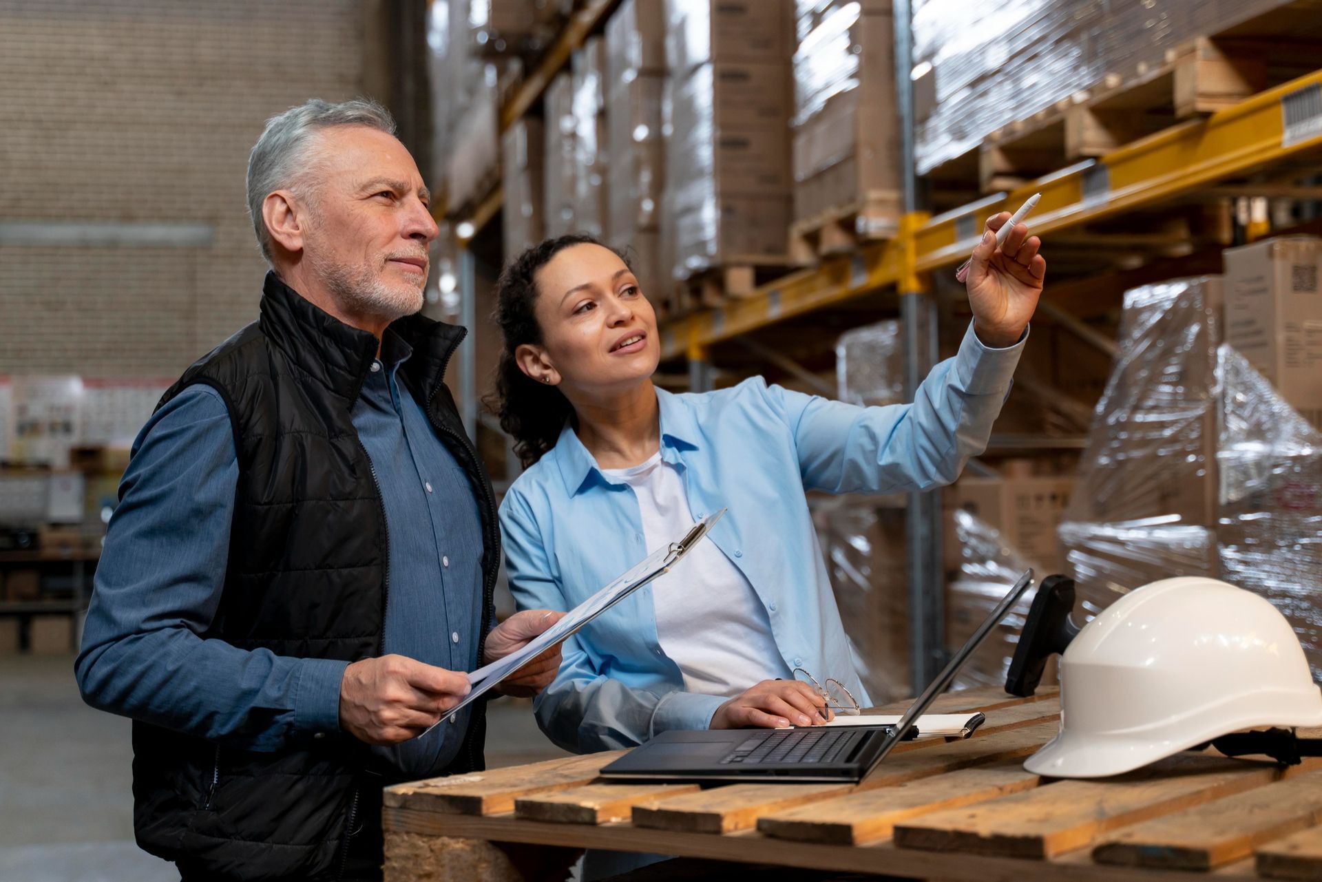 Man and woman in warehouse, inspecting inventory, woman pointing, notebook, hard hat on pallet.