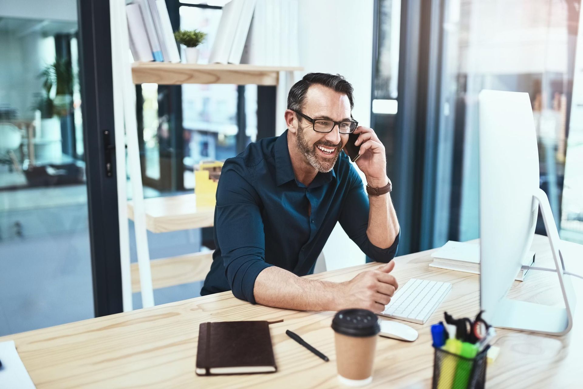 Man in glasses smiles while on the phone at a desk with a computer, coffee, and notebook.