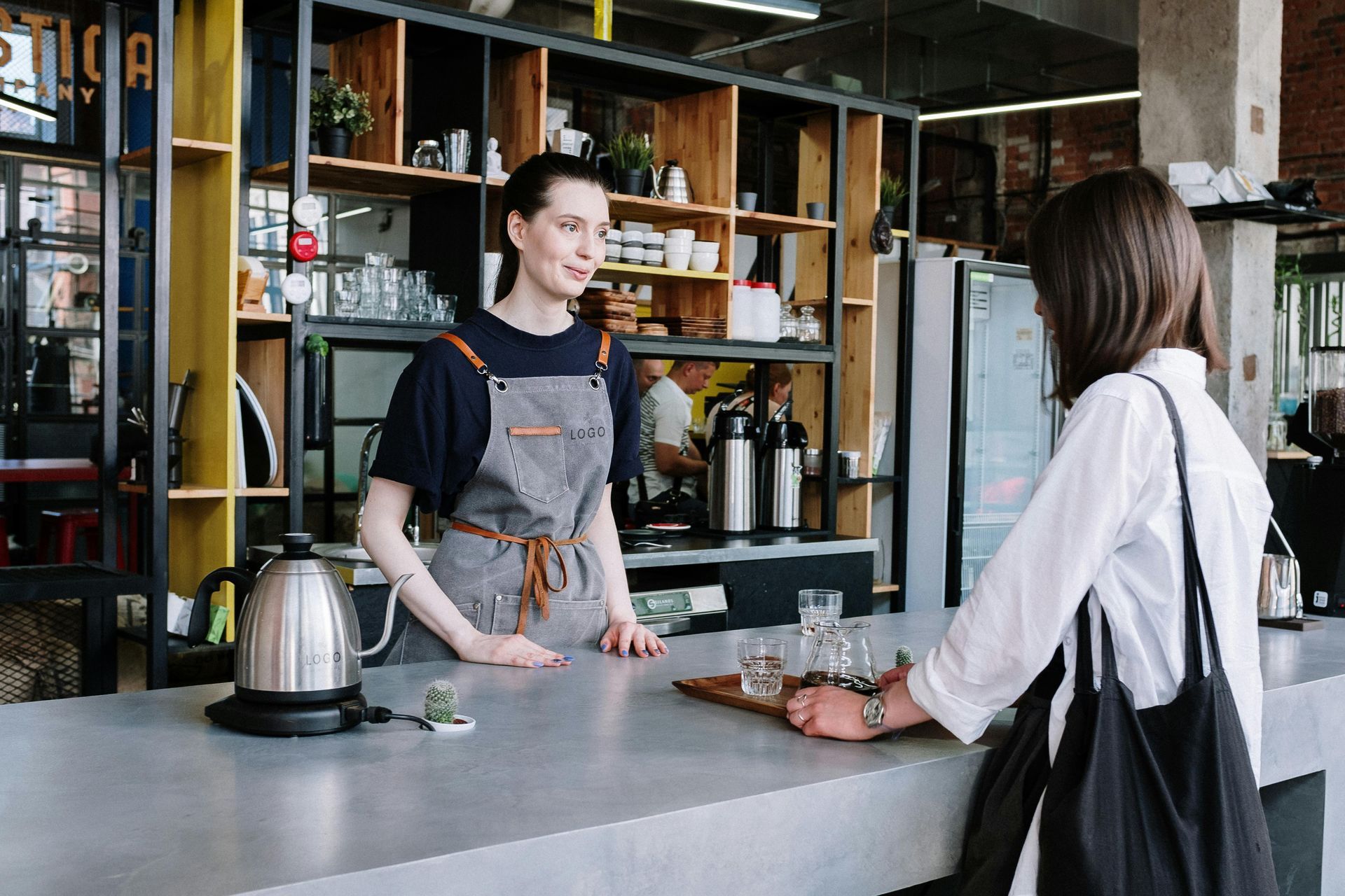 Woman barista taking an order at a cafe counter. A customer stands facing her, with glasses and a tray.