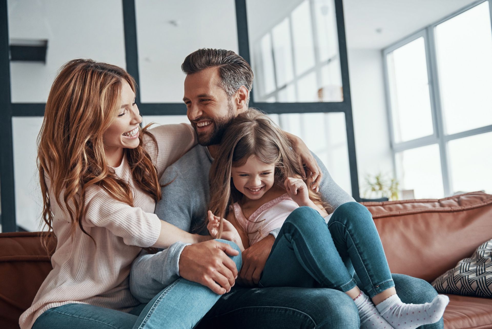 Family of three laughing on a couch. Living room with large windows, neutral tones.