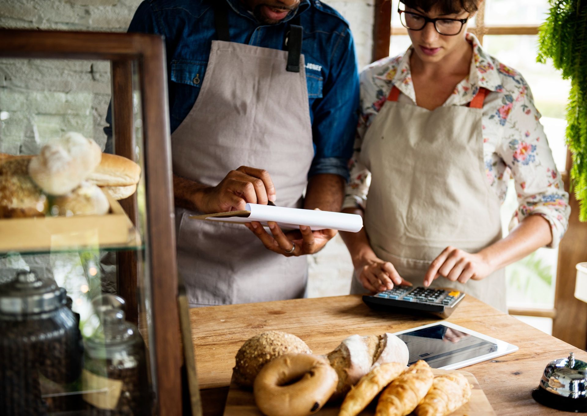 Two people in aprons calculate bakery sales with clipboard and calculator at counter.