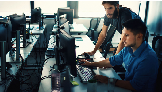 Two men working at computers in an office setting, one looking at the screen, the other leaning in and observing.