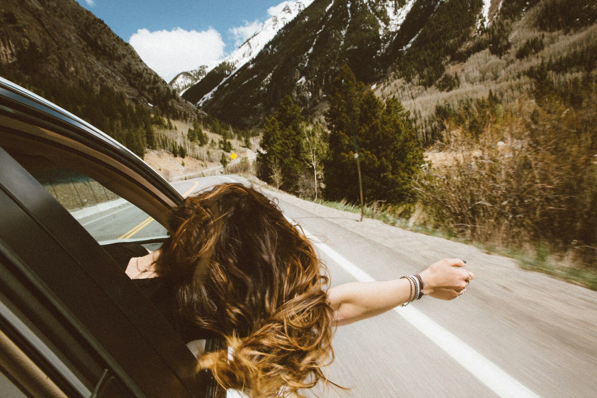 Woman leans out of car window, arm outstretched, driving on a mountain road with snow-capped peaks in background.