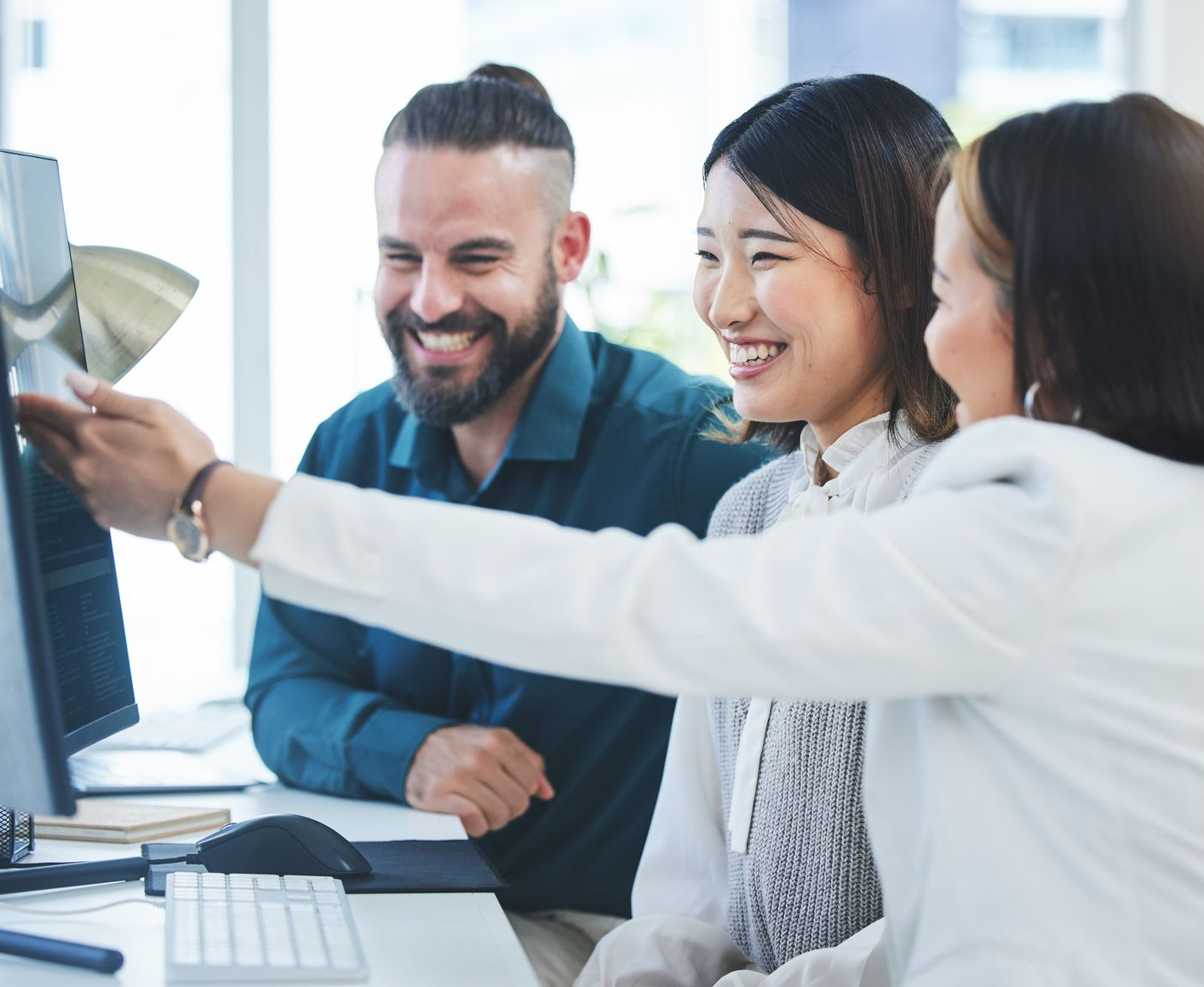Three people smiling, looking at a computer screen in an office setting. One points at the screen.