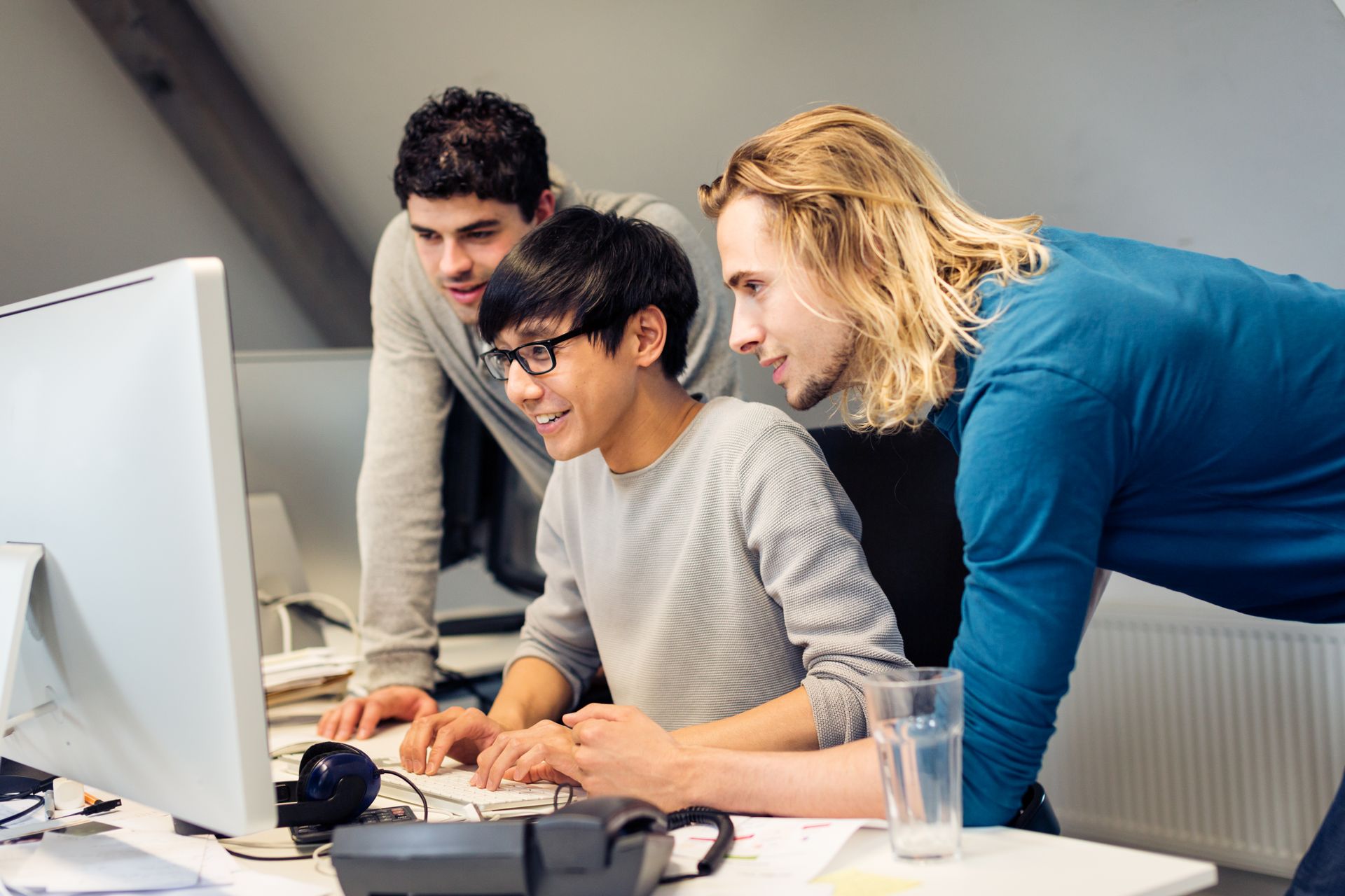 Three people look at a computer screen, one typing, others leaning in. Bright office with papers.
