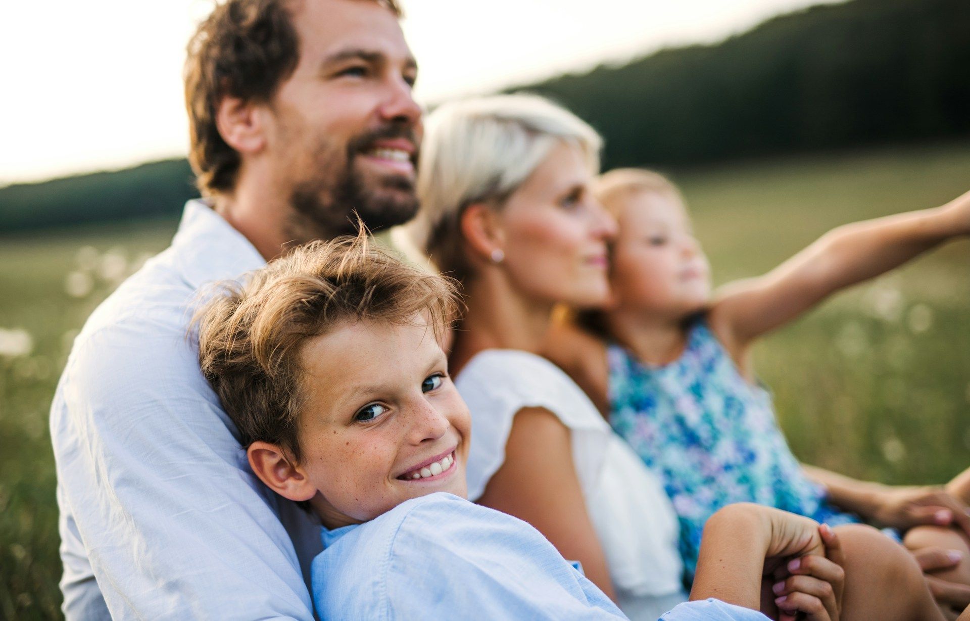 Family of four sitting outdoors, looking up and smiling. Boy in front smiles at camera.