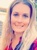 Blonde woman with blue eyes smiles outdoors, wearing a red plaid shirt, blurred background.