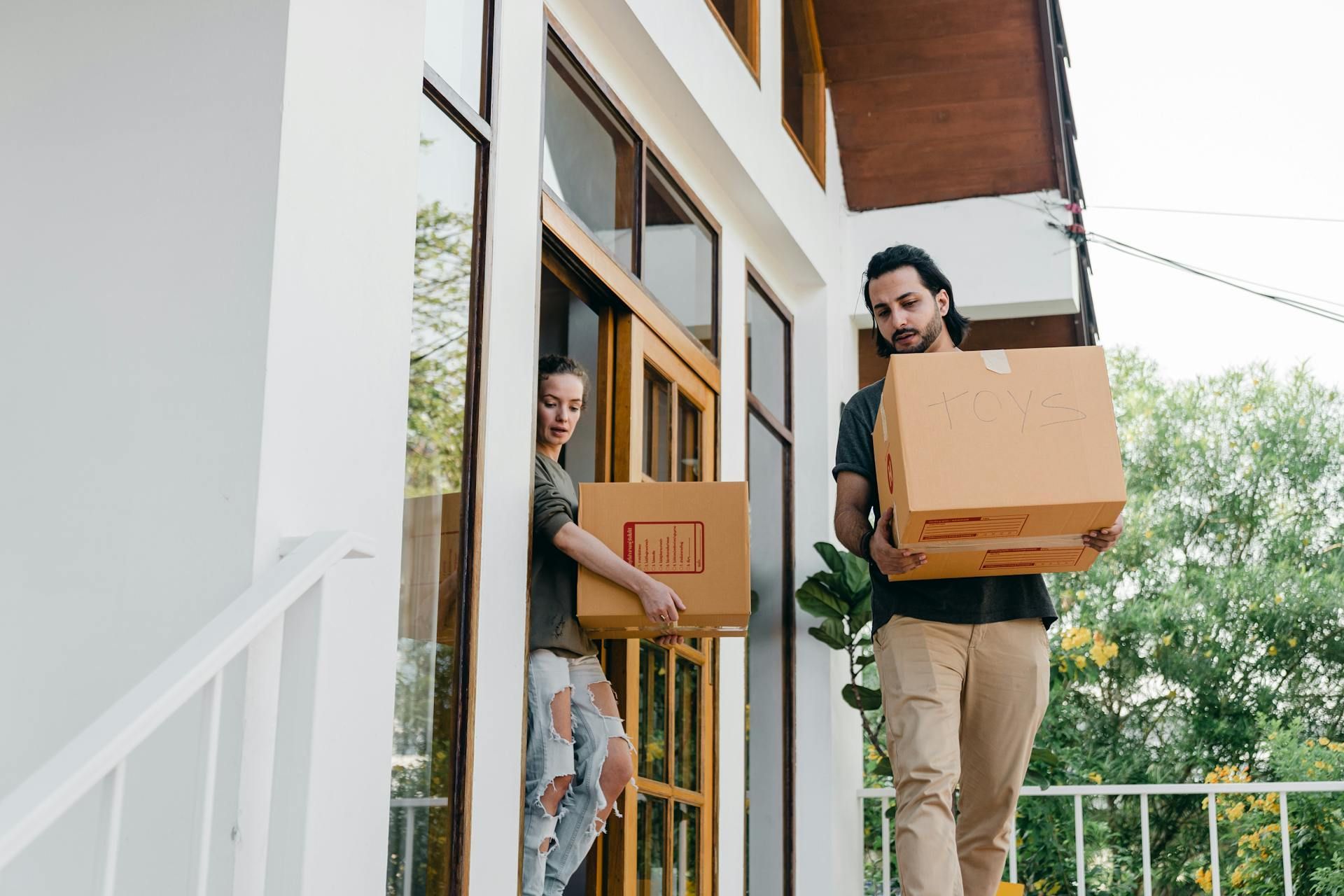 Couple carrying moving boxes out of a white house with large windows.
