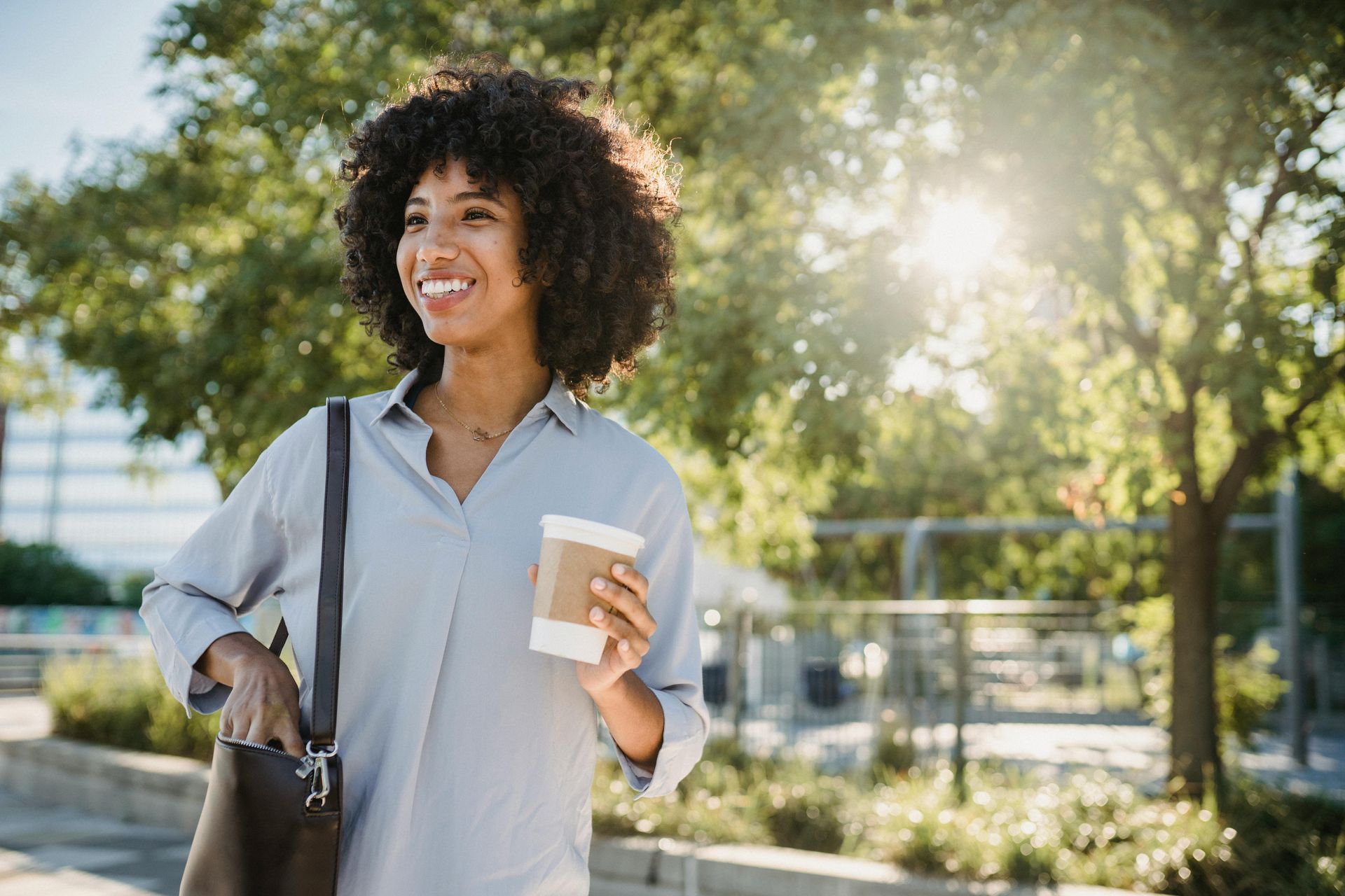 Woman with coffee cup, smiling outdoors, sunlight, near trees.
