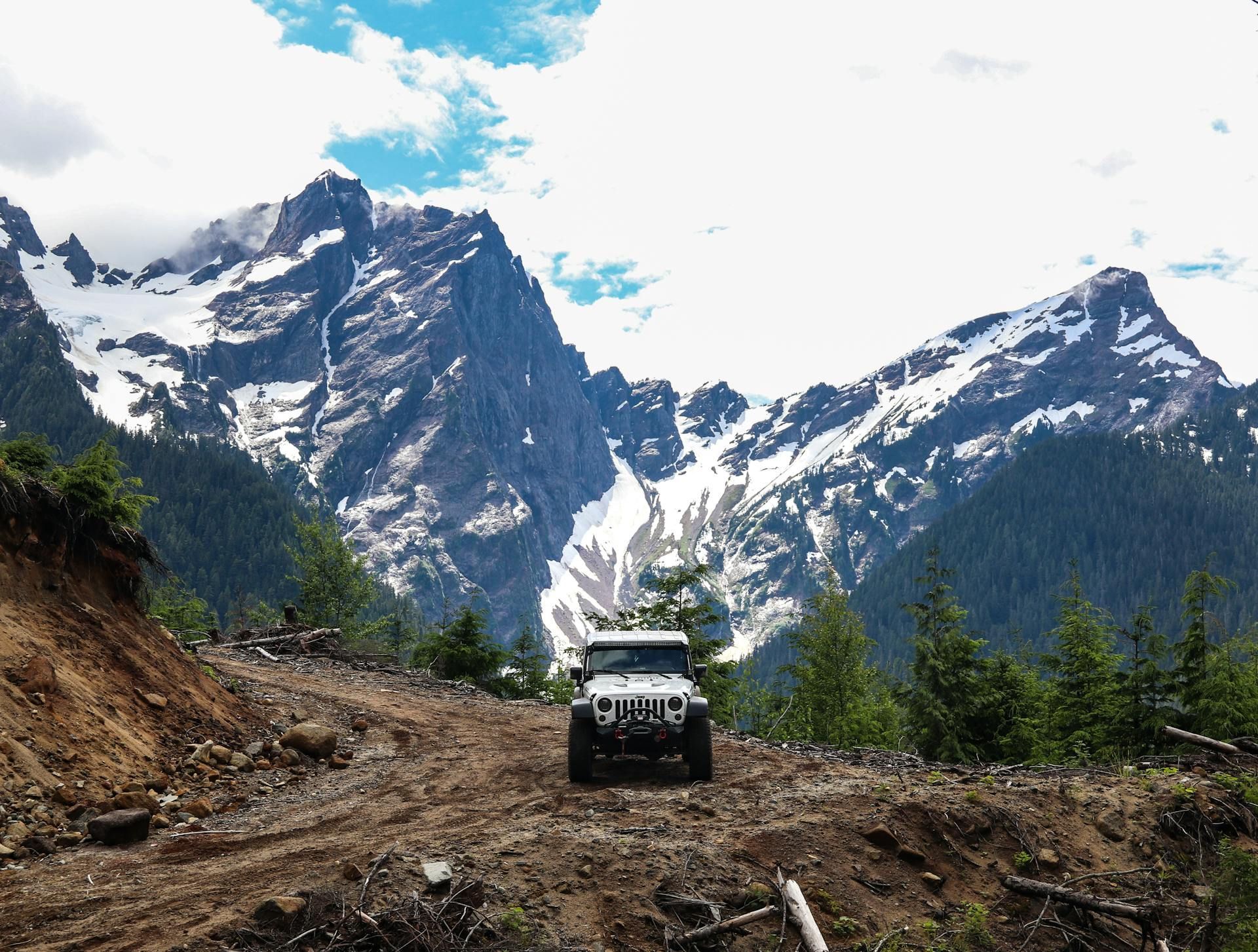 White Jeep on a dirt road in mountains with snow. Cloudy sky and green trees.