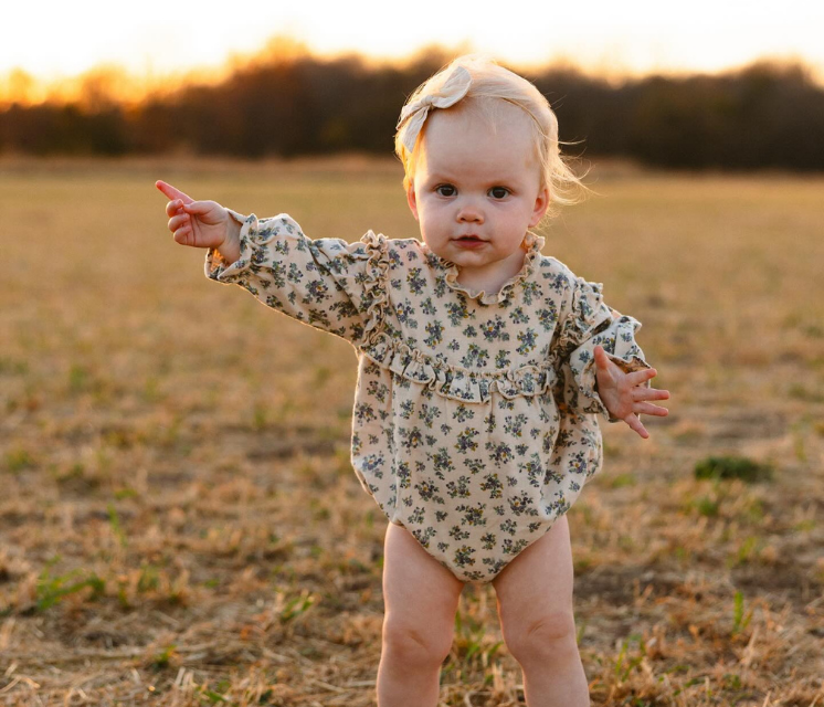 Blonde baby in floral onesie points towards the golden sunset in a field.