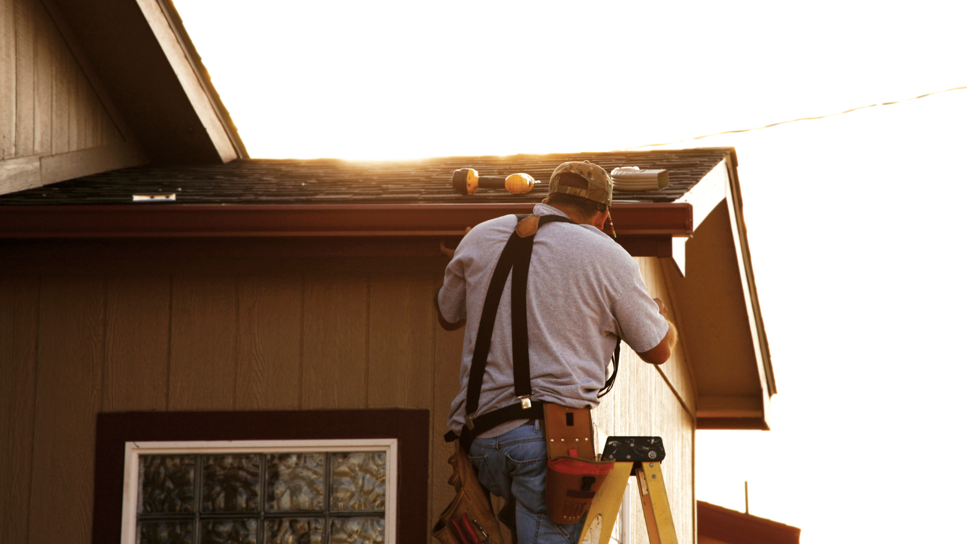 Man on ladder installing gutter on a house, back to camera, sunlit.