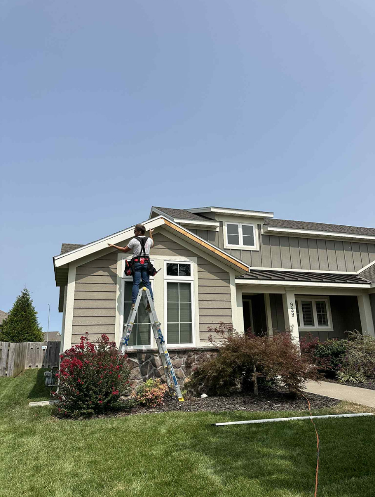 Person on a ladder painting house trim. Tan siding, blue sky, green grass.