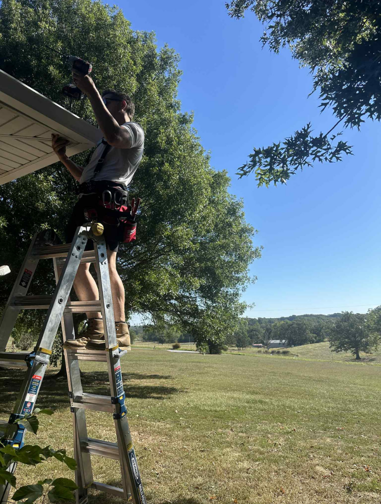 Man on ladder, drilling something under the roof on a sunny day. Green trees and field in the background.