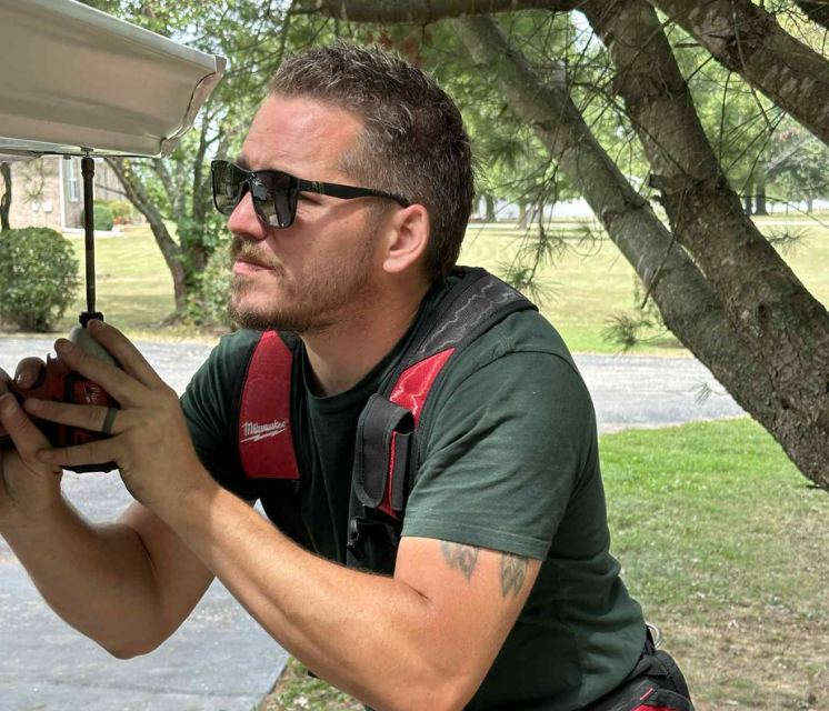 Man in sunglasses working, wearing a red tool harness, working outside near a tree.