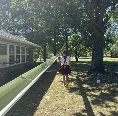 Person carrying a long green gutter on a grassy path near a brick building and trees on a sunny day.