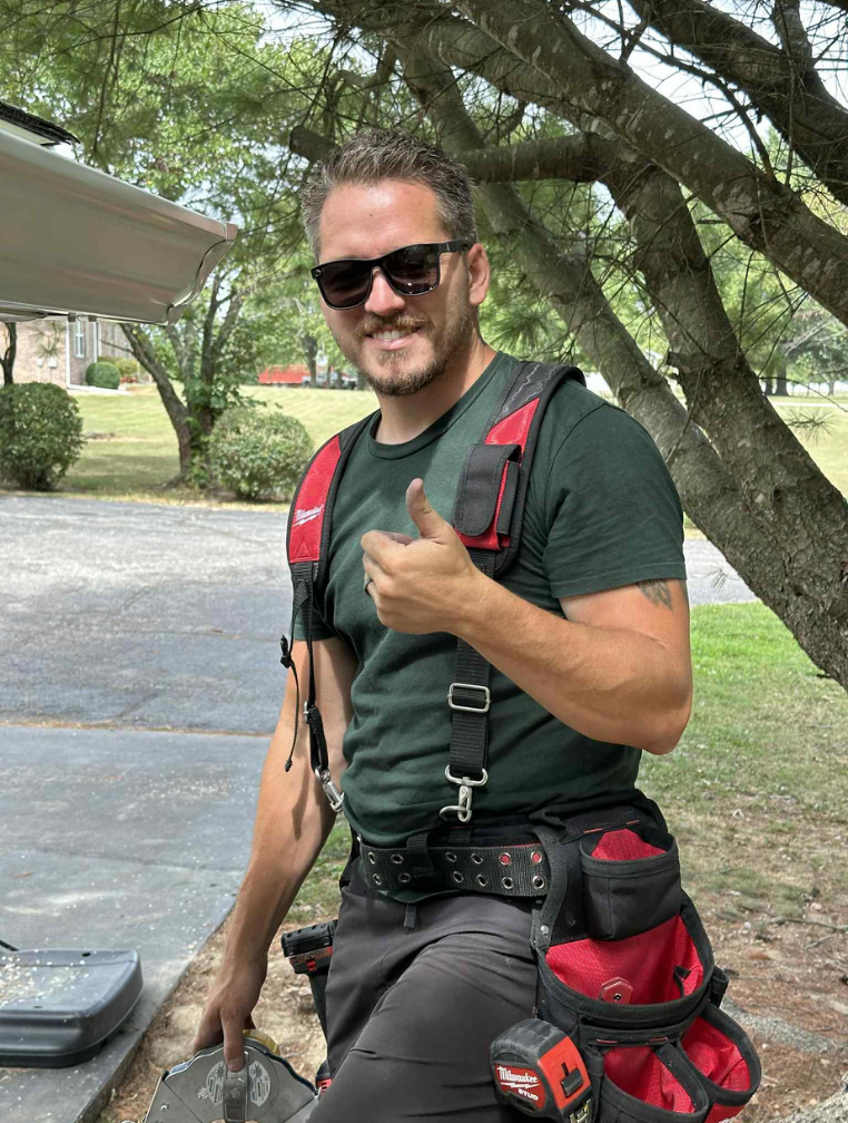 Man in sunglasses gives thumbs up, wearing tool belt in a yard.