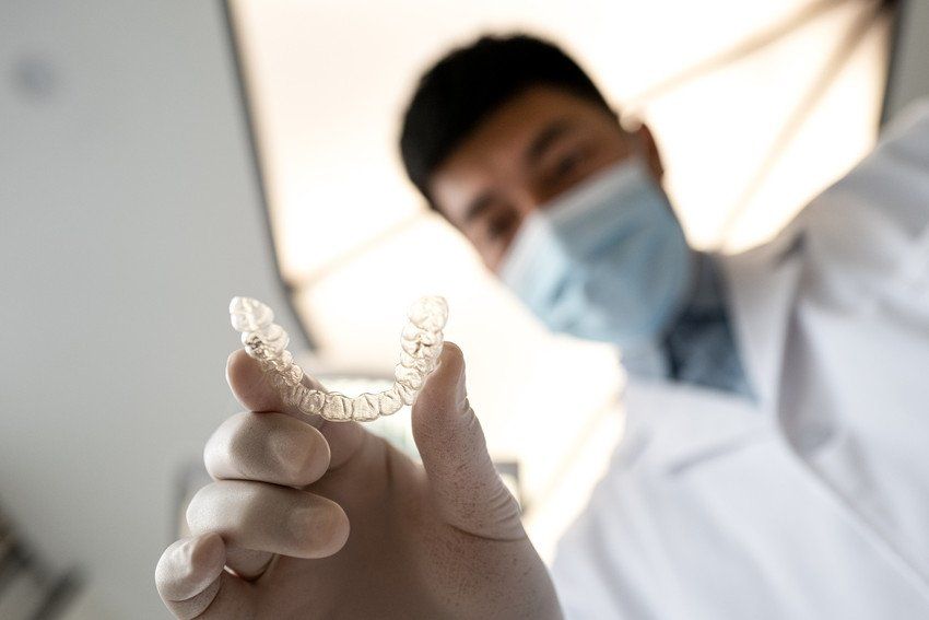 A dentist wearing a mask and gloves is holding a clear brace in his hand.