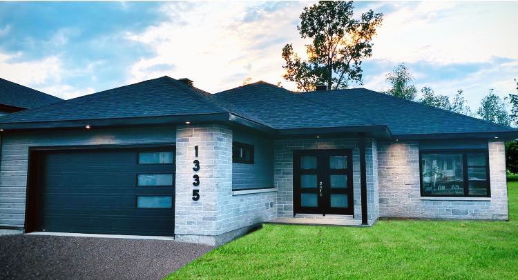 Maison moderne de plain-pied avec façade en briques grises, porte de garage noire et toit sombre. Porte d'entrée noire avec vitres. Pelouse verte.