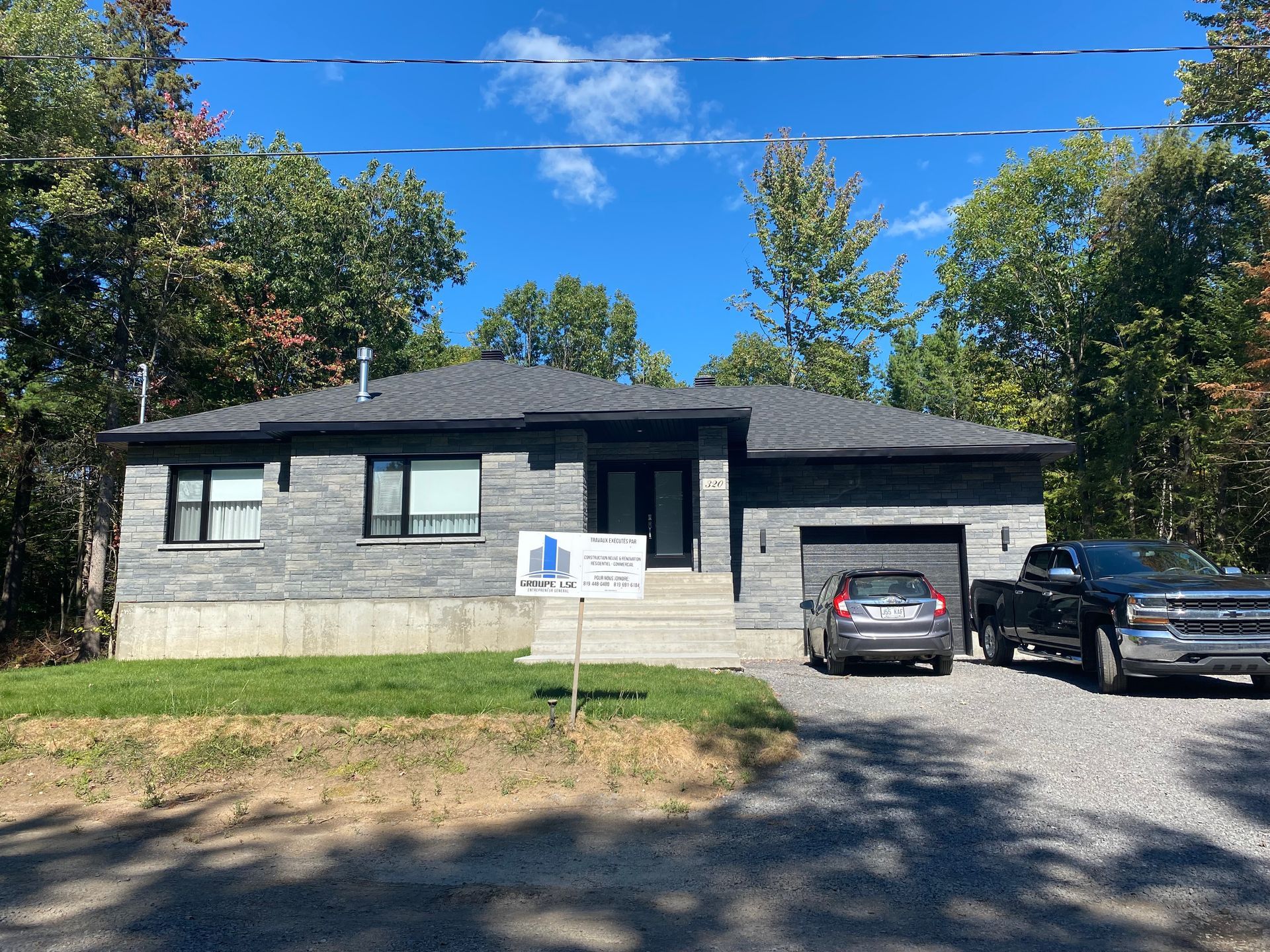 Maison moderne en pierre grise avec un garage et des voitures garées dans l'allée, sur fond d'arbres verts sous un ciel bleu.