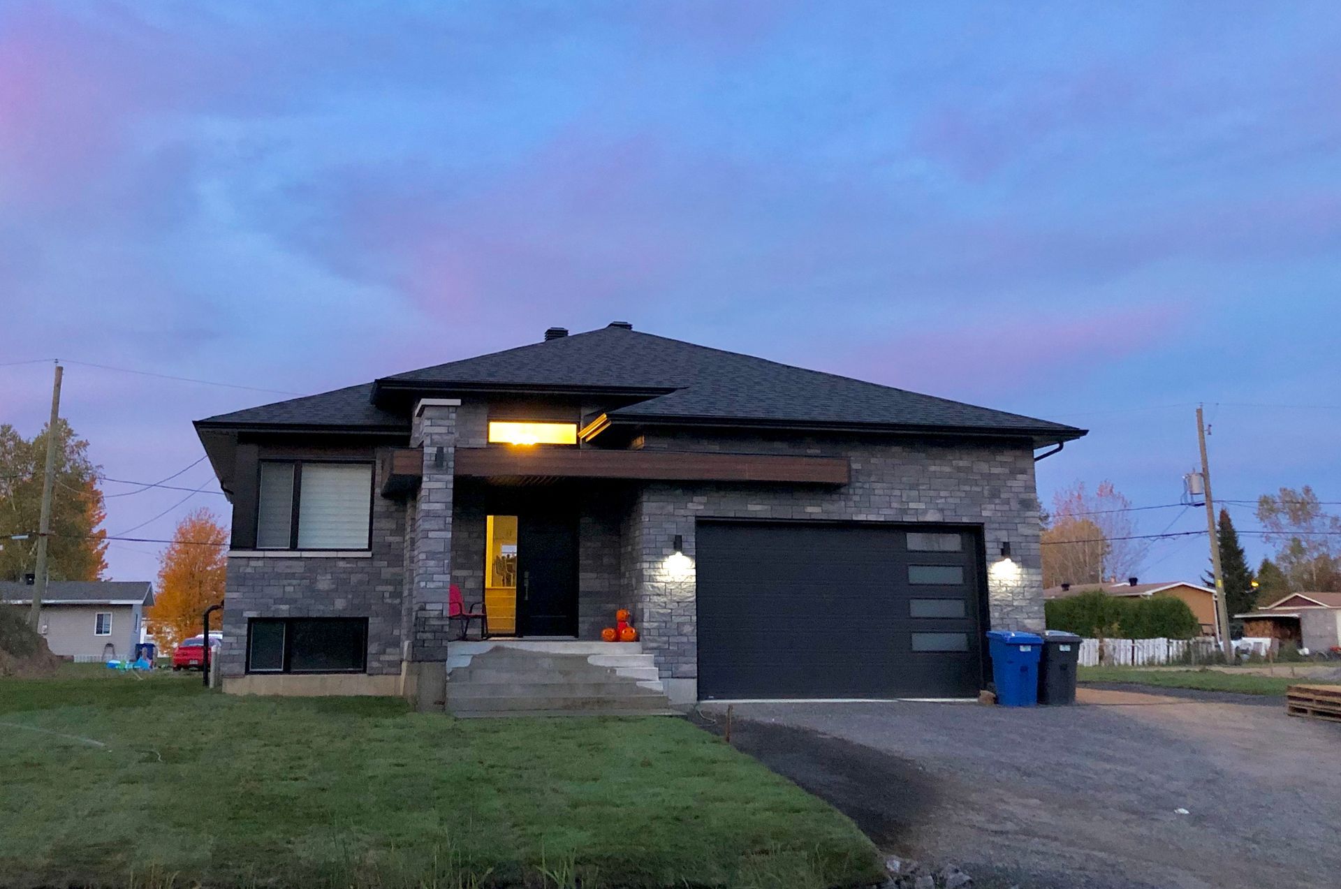 Maison moderne avec façade en pierre grise et porte de garage gris foncé au crépuscule. Le ciel est teinté de rose et de bleu.