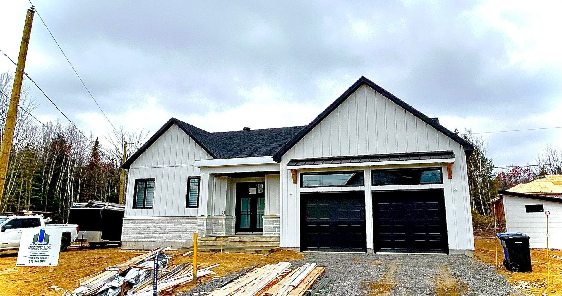 Une nouvelle maison en construction, avec un revêtement blanc, des portes de garage noires et un toit sombre, contre un ciel nuageux.