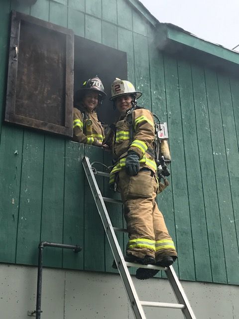 Two firefighters are standing on a ladder looking out of a window.