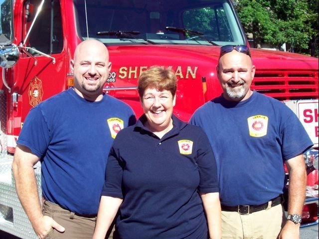 Three people standing in front of a red truck that says shefman