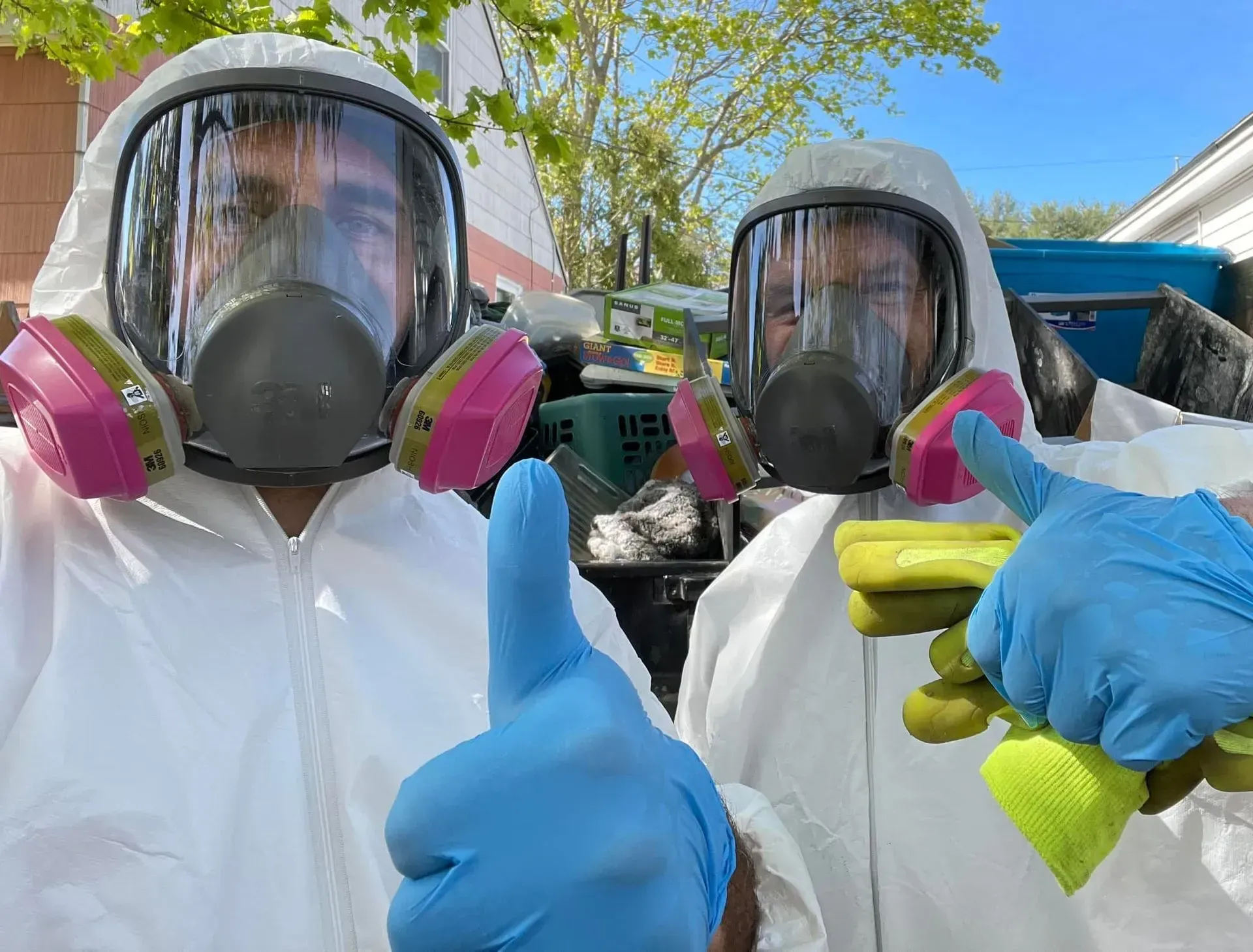 Two men wearing gas masks and gloves are giving a thumbs up.