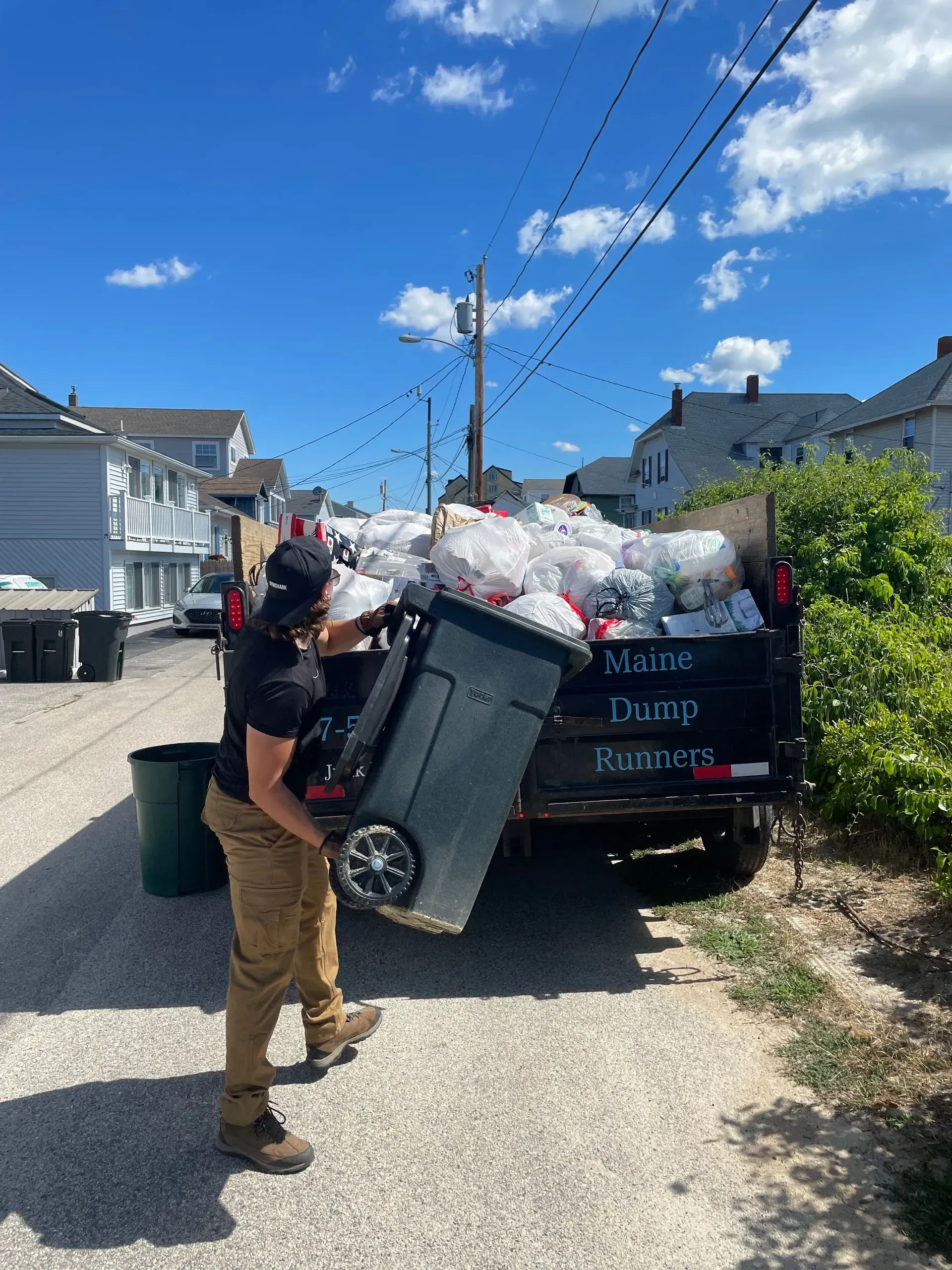 A man is pushing a garbage can in front of a dumpster filled with trash.