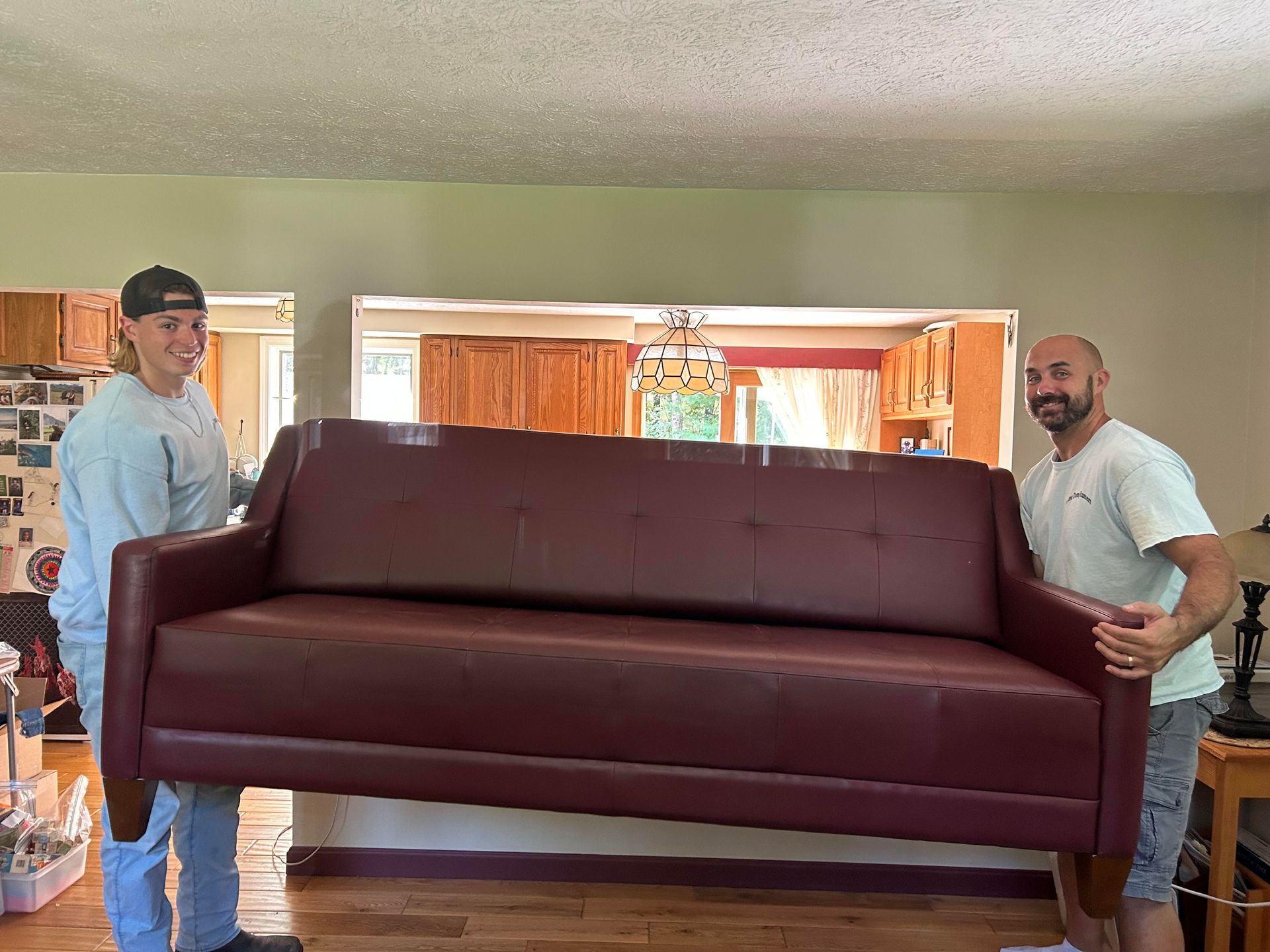 Two men are carrying a large red leather couch in a living room.
