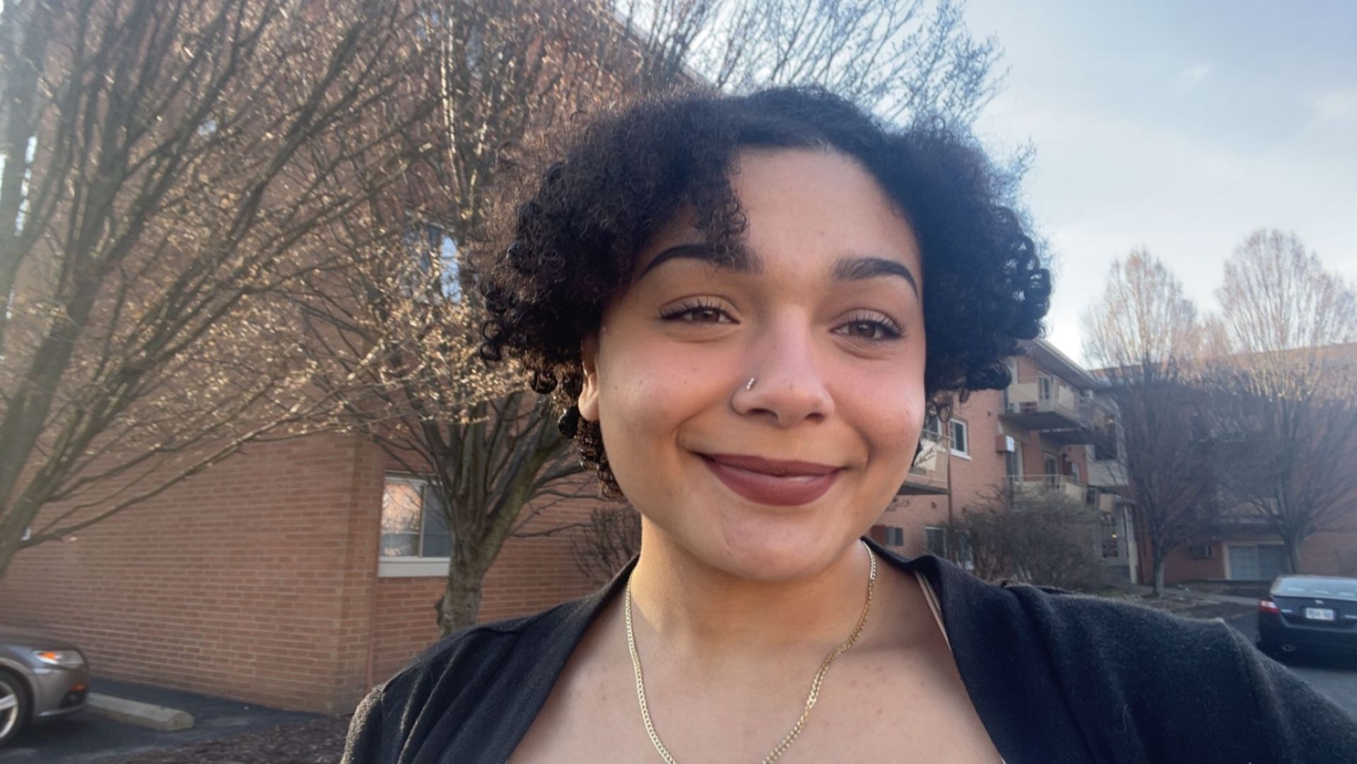 A woman with curly hair and a nose ring is smiling in front of a brick building.