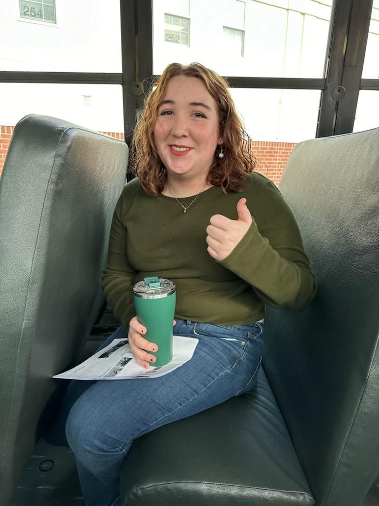 A woman is sitting on a bus holding a green cup and giving a thumbs up.