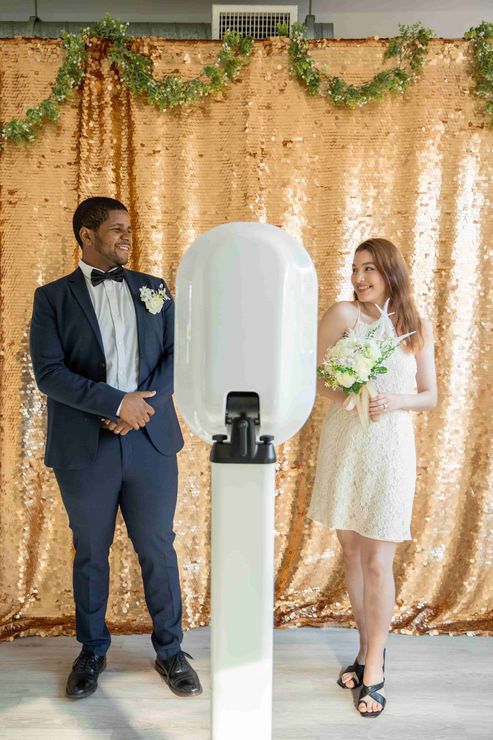 One man and one woman dressed for a wedding standing in front of a digital photo booth with a golden backdrop