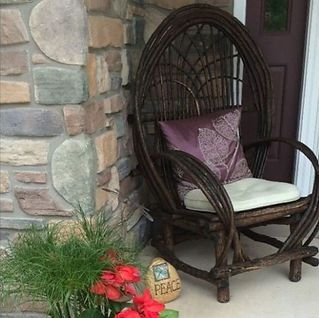 A wicker chair with a purple pillow is sitting on a porch next to a stone wall.