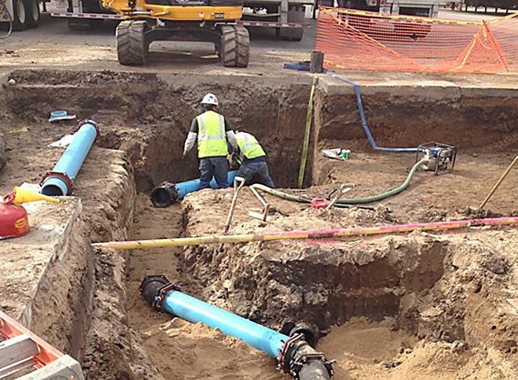 A man in a yellow vest is working on a pipe in the dirt