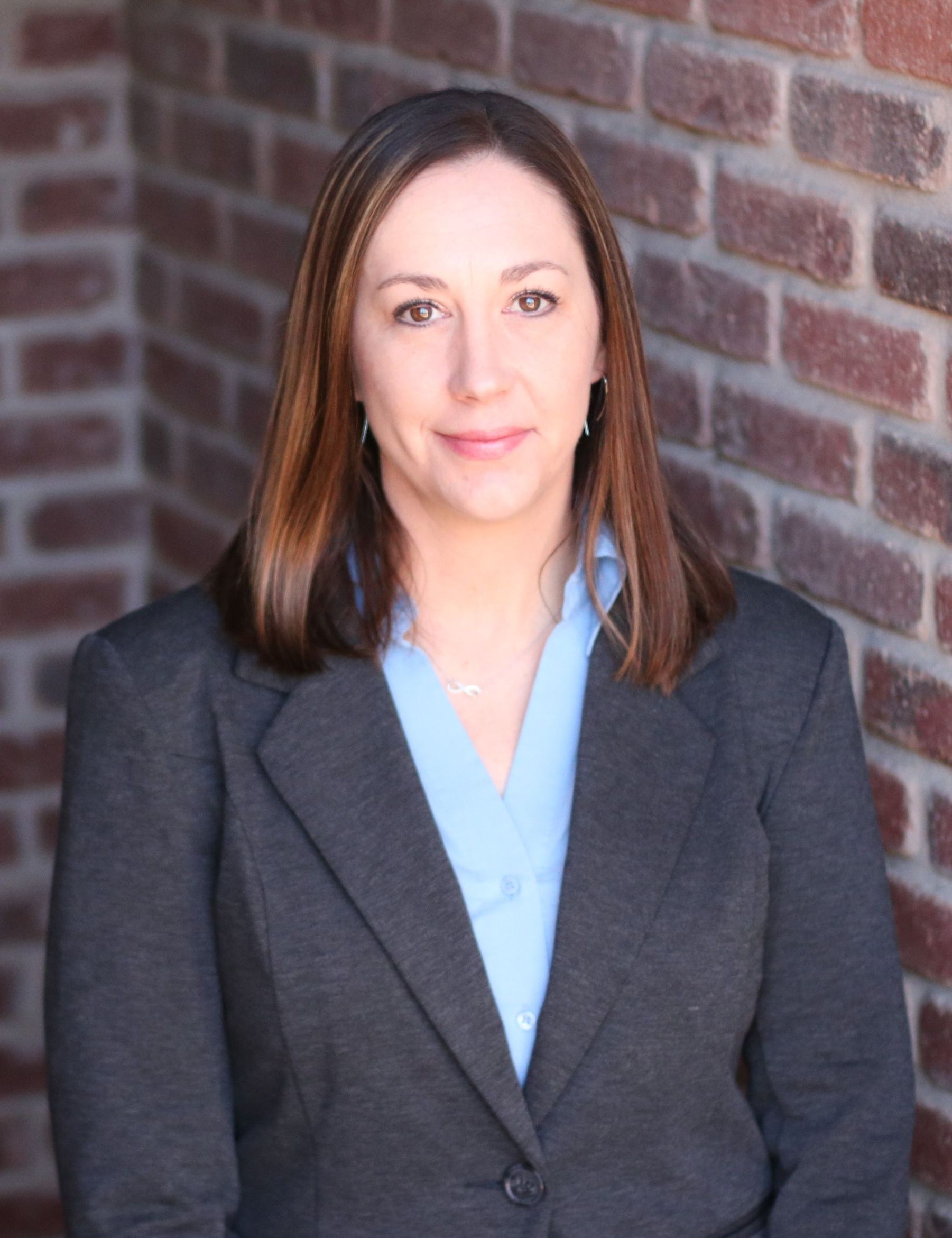 A woman named Kayla Leanos in a suit and blue shirt is standing in front of a brick wall.