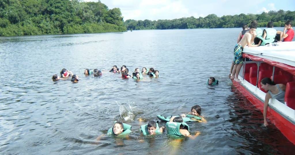 Personas nadando en un lago cerca de un bote rojo y blanco, algunos subiéndose. Árboles al fondo.