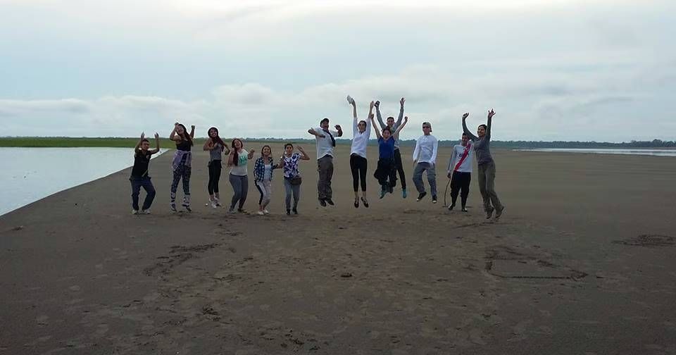 Grupo de personas saltando en un terraplén de arena cerca del agua, con los brazos levantados en celebración.