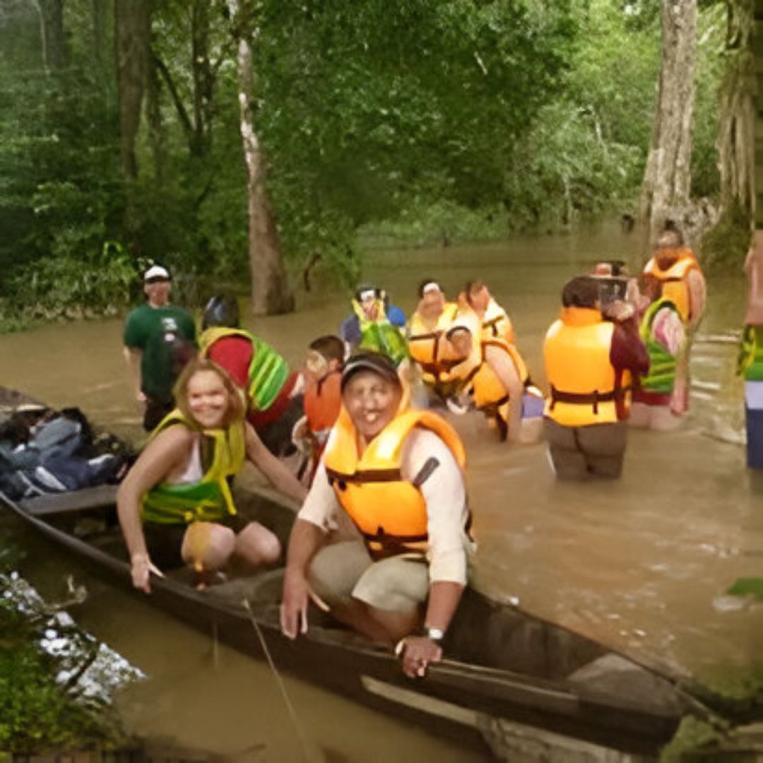 Personas con chalecos salvavidas en un bote en un río. Árboles verdes al fondo.