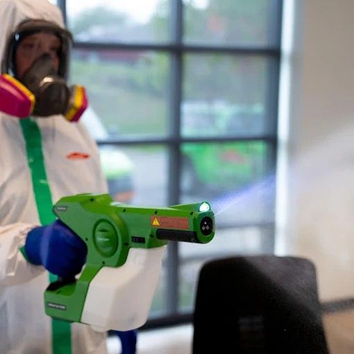 Person in protective suit using a green disinfectant sprayer.
