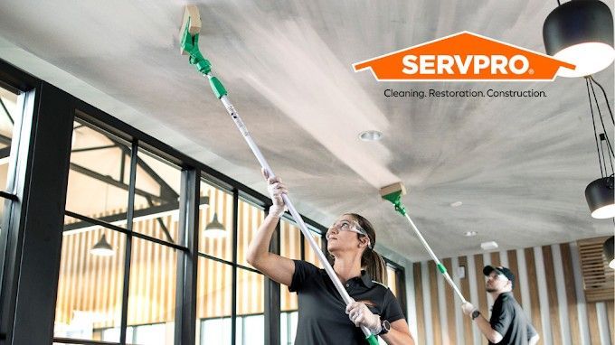 Two people cleaning a ceiling with spray wands in a commercial space. SERVPRO logo visible.