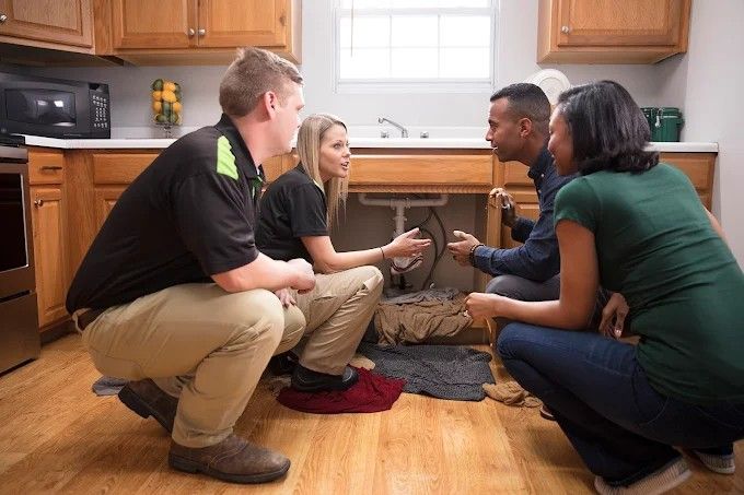 Two professionals discuss plumbing issues with a couple in a kitchen.