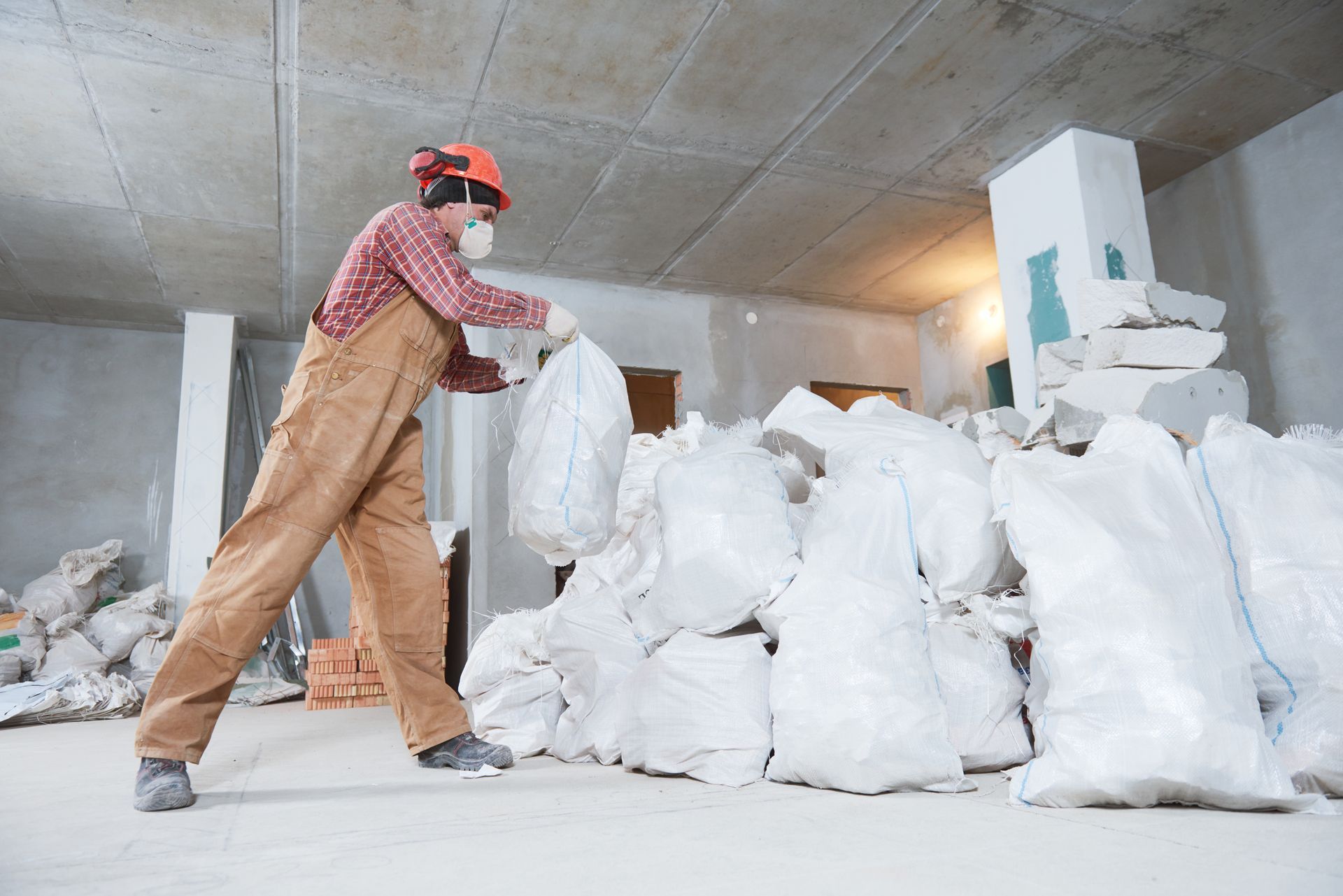 Person in overalls and respirator bags construction debris in a room with a concrete ceiling.