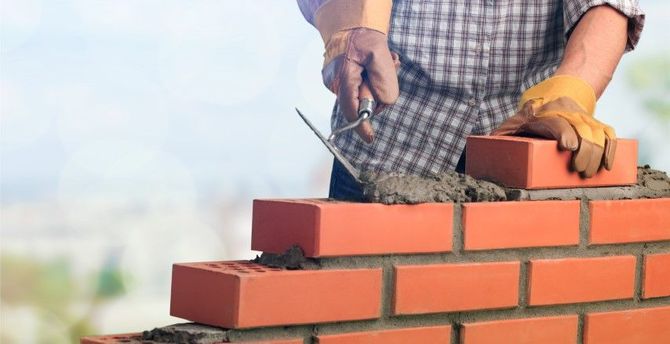 Worker laying red bricks with mortar on a brick wall, wearing orange gloves outdoors