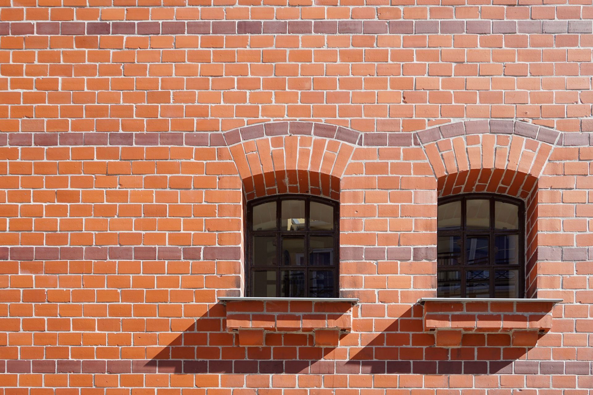Brick wall with two arched windows and white trim, lit by sunlight and casting shadows