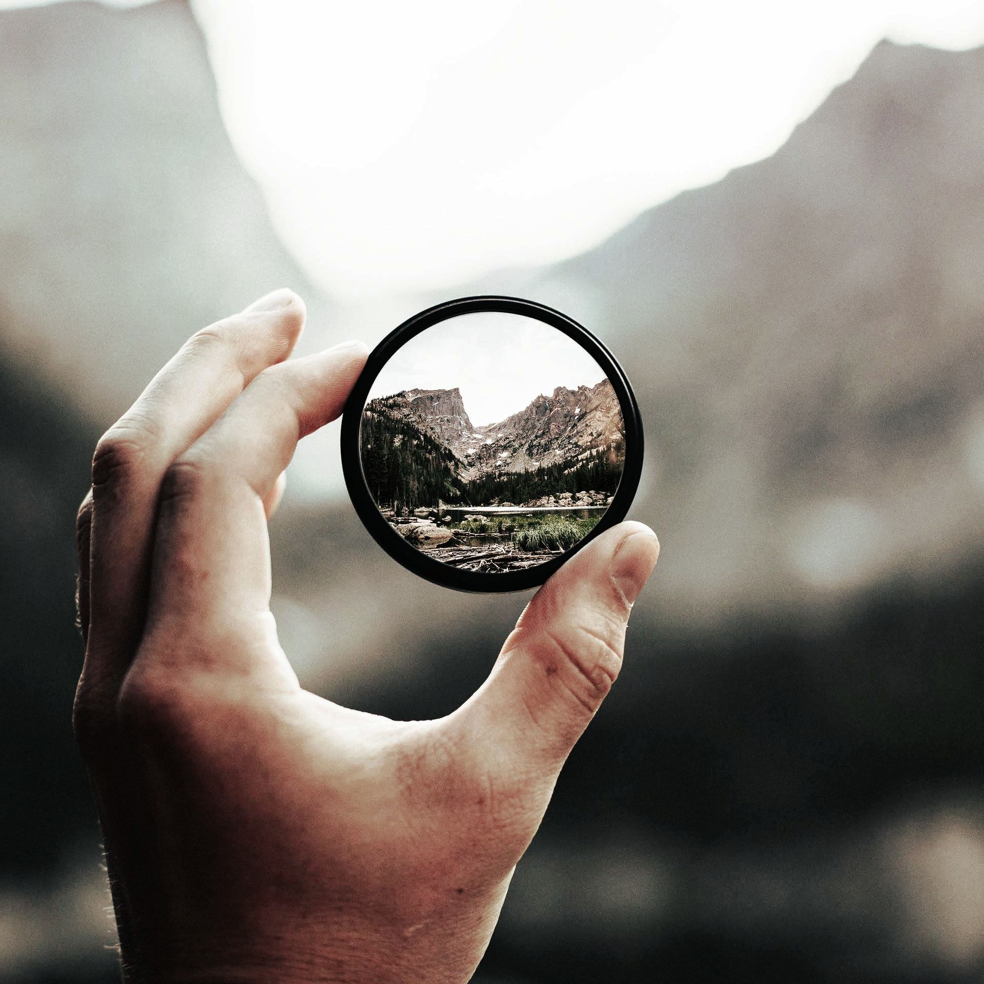 Hand hält eine runde Linse, fokussiert auf Berge und See; unscharfer Hintergrund.